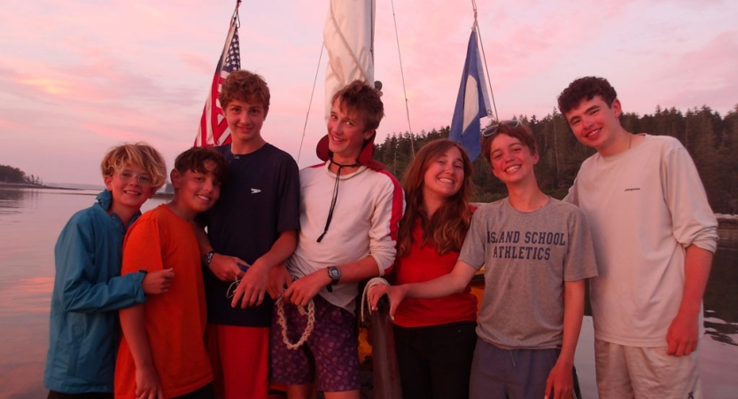 A group of students gather close together for the photo. They are near a body of water. 